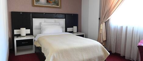 Habitación de hotel con una cama grande, una mesita de noche con lámpara, un cuadro en la pared y una ventana con cortinas.