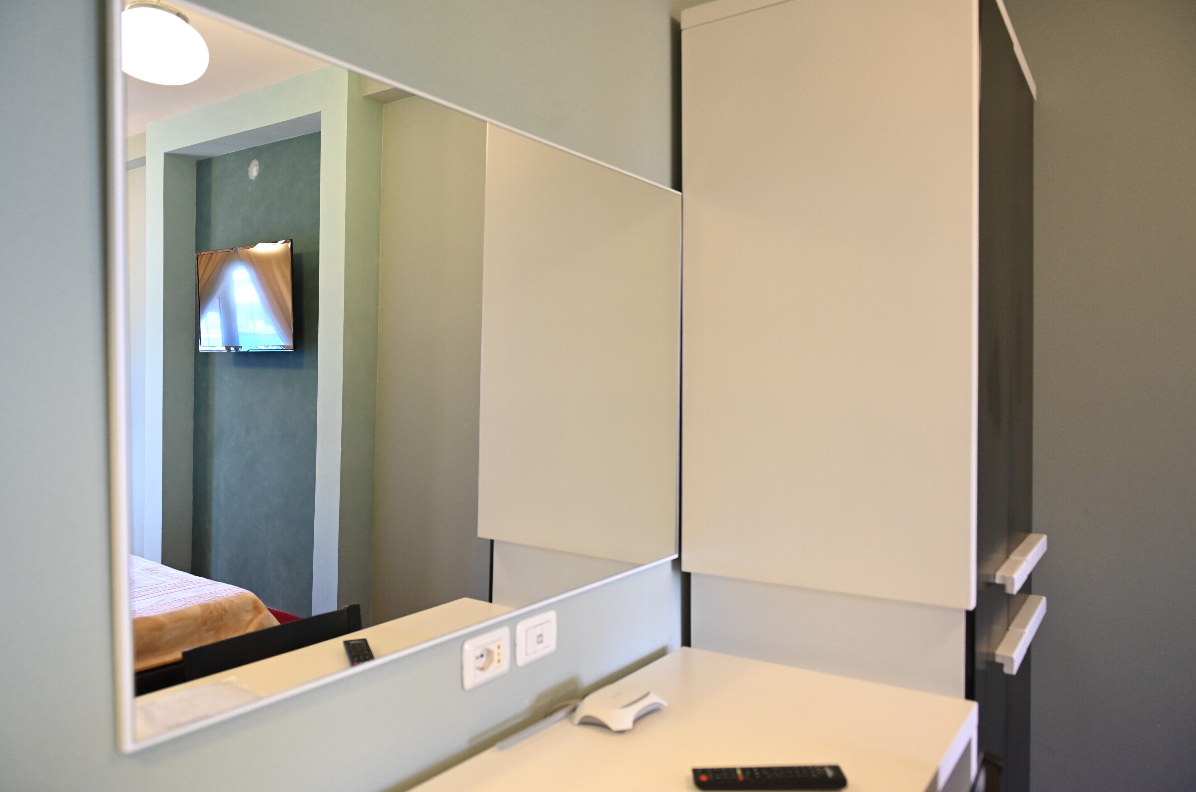 A hotel room with a large mirror, a bedside table with a phone, and a small window with curtains.