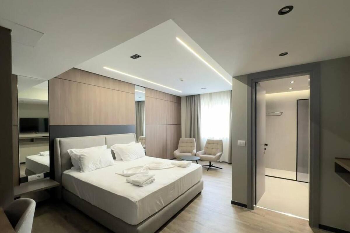 A modern hotel room with a large bed, a seating area with two chairs, and a walk-in closet.