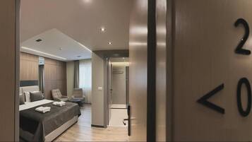 A modern hotel room with a bed, two armchairs, a desk, and a door with a number sign.