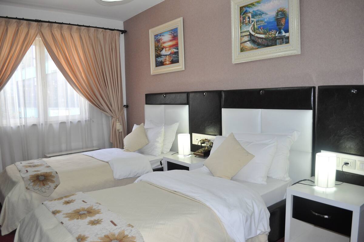 A hotel room with two beds, a nightstand with a lamp, and two framed pictures on the wall.