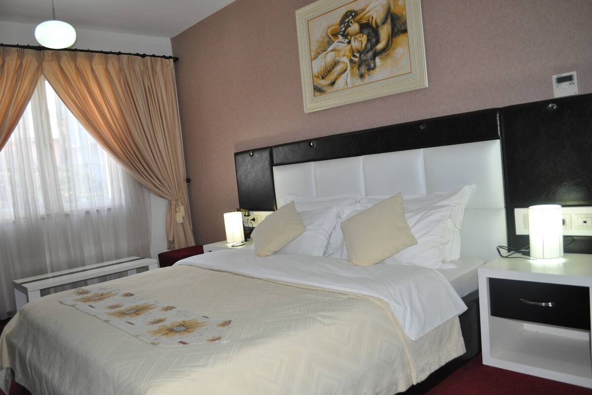 A hotel room with a large bed, a nightstand with a lamp, a wall-mounted painting, and a window with curtains.