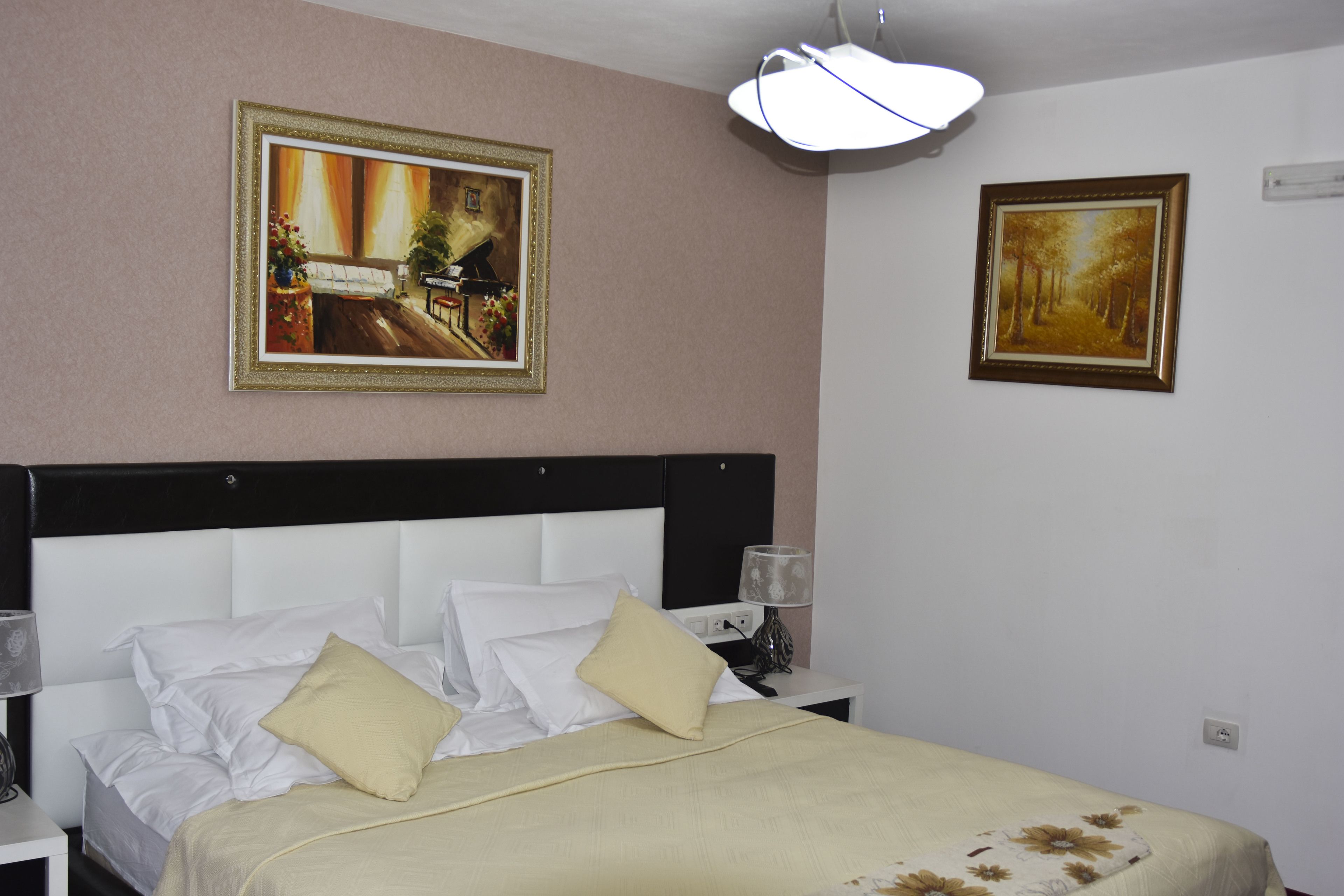 A neatly made bed with white and beige bedding, two bedside lamps, and two framed paintings on the wall.