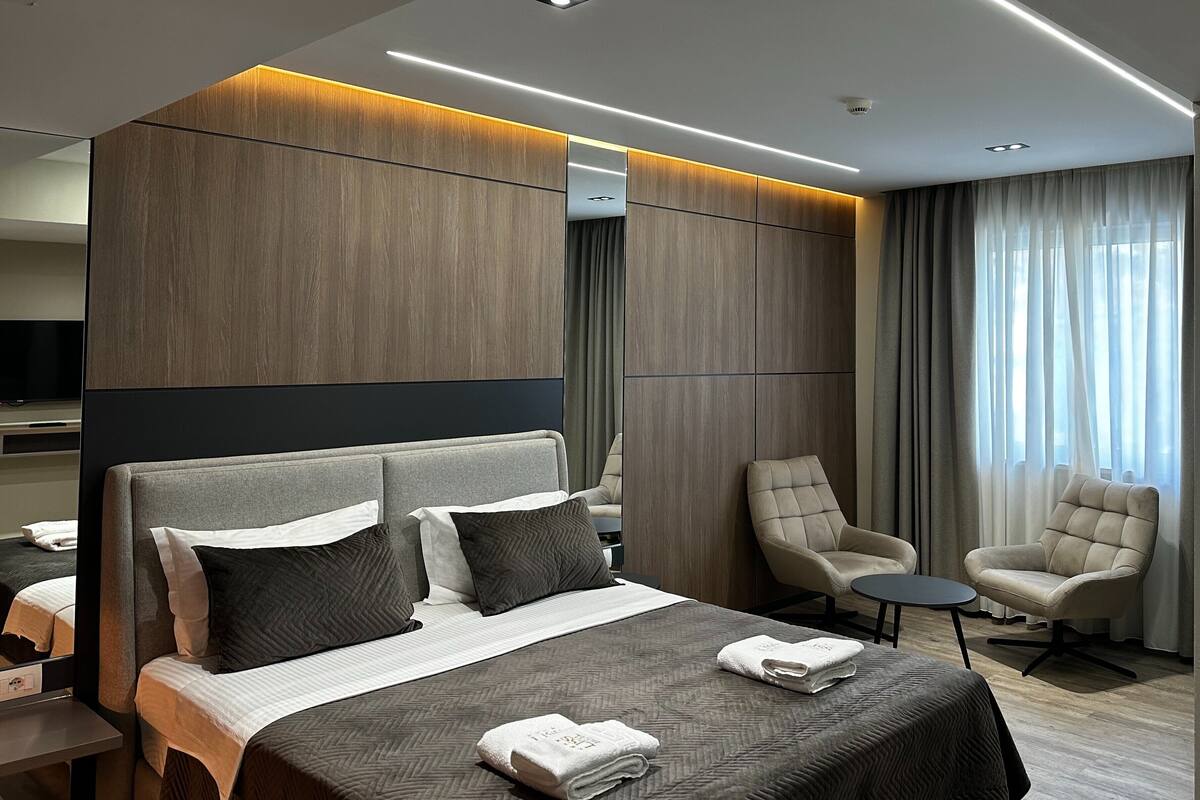 A modern hotel room with a large bed, two armchairs, a small table, and a television.