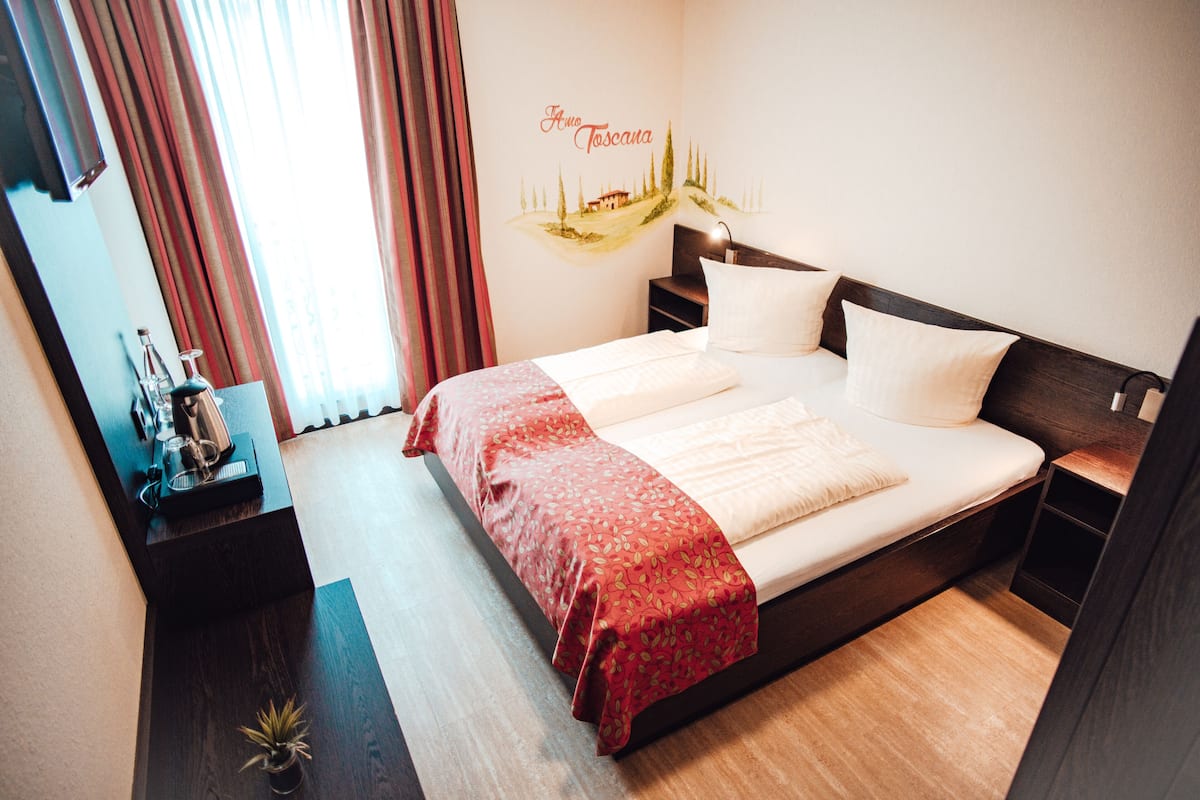 A neatly made bed with a red and white bedspread, a bedside table with a lamp, and a wall decoration depicting a Tuscan landscape.