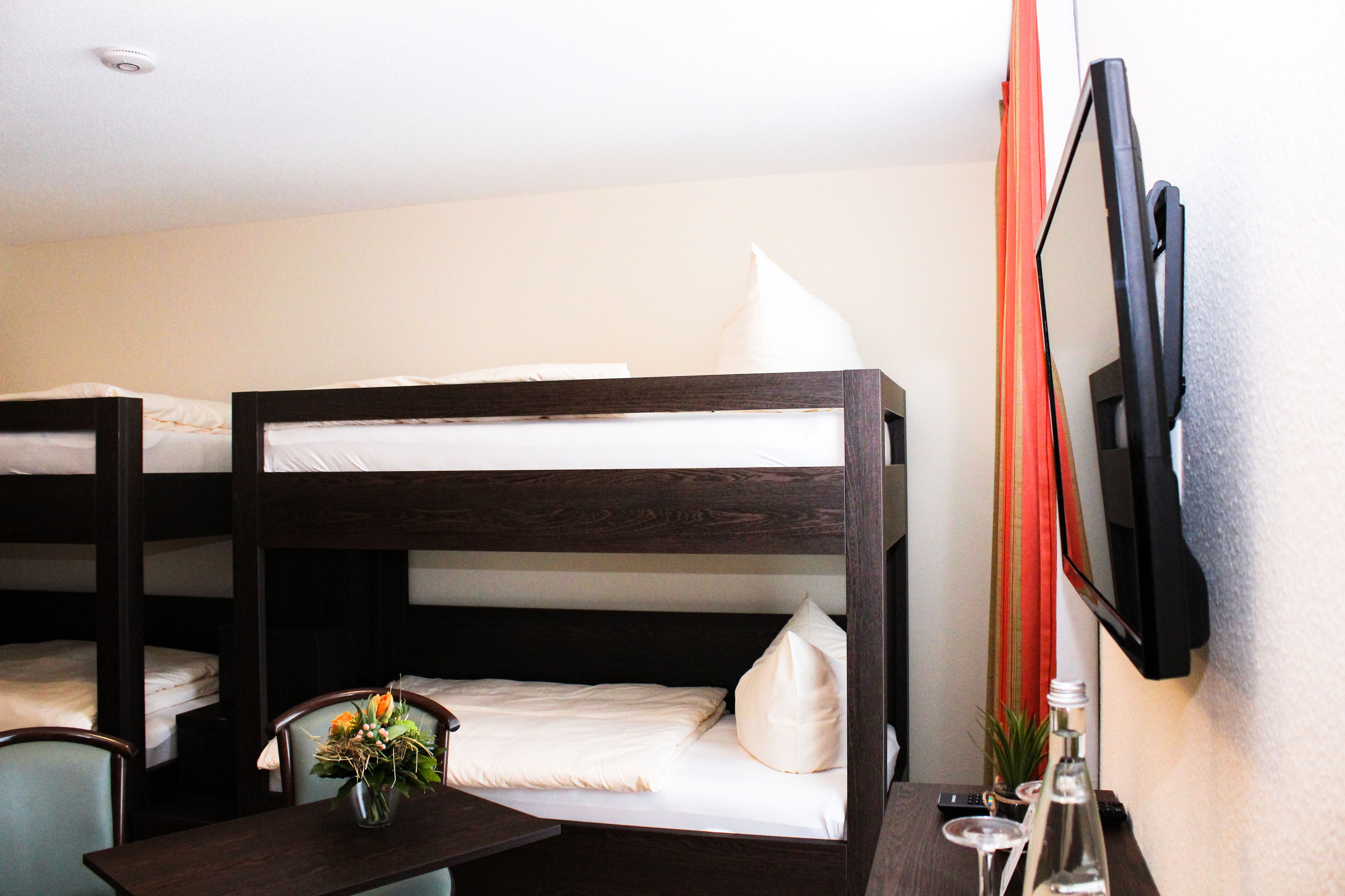 A bunk bed room with a desk and chair, a TV mounted on the wall, and a small table with a vase of flowers.