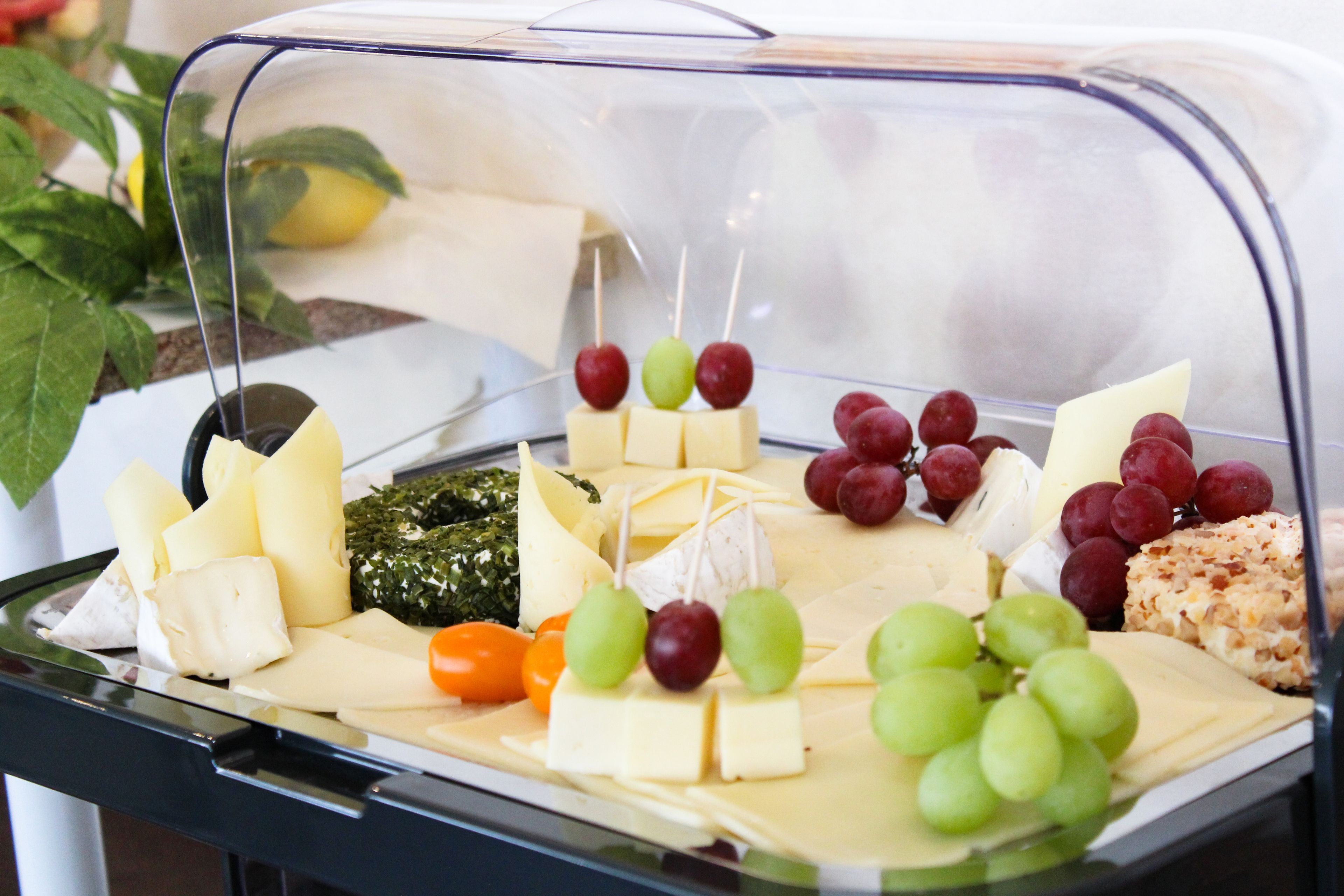 A cheese and fruit platter with various cheeses and grapes displayed in a clear plastic container.