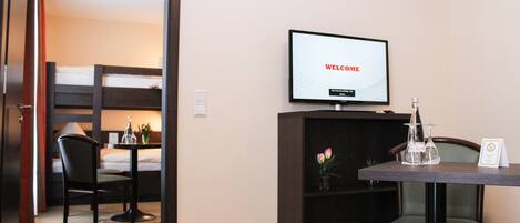 Quarto de hotel com cama, escrivaninha com monitor de computador exibindo “WELCOME”, cadeira, mesinha de centro com um vaso de flores e televisão fixada na parede.