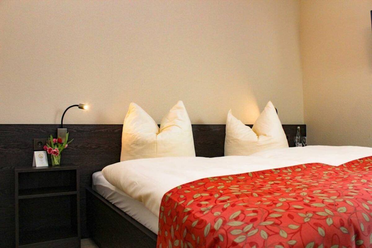 A hotel room with a bed, white pillows, a red patterned bedspread, a nightstand with a lamp, and a vase with flowers.