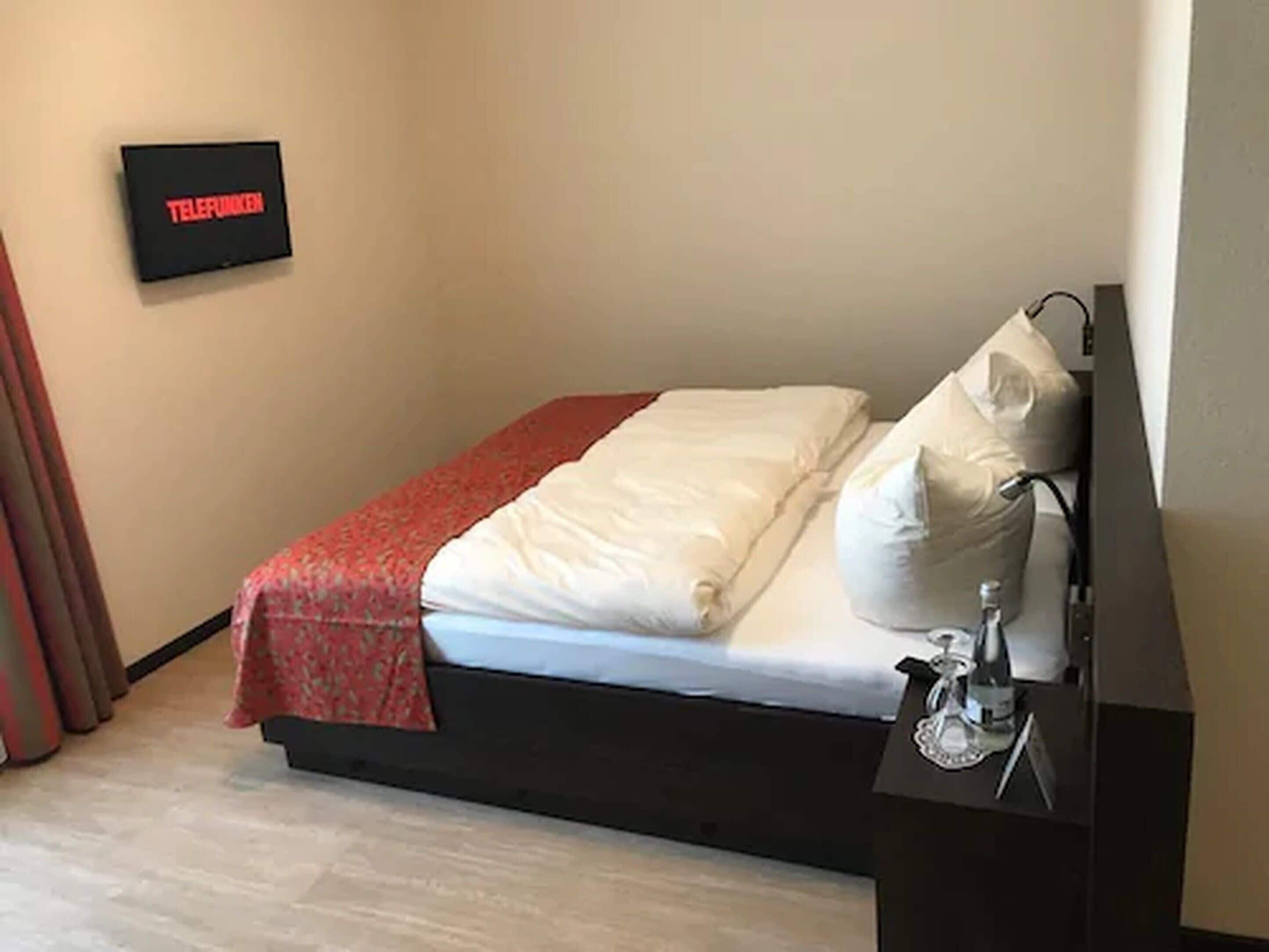 A hotel room with a bed, a bedside table with a lamp, and a wall-mounted TV displaying 'TELEFUNKEN'.