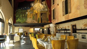 A restaurant with yellow chairs, tables set with white tablecloths, and a mural of a village on the wall. - Hotel La Toscana Nähe Europapark  mit Ticket Garantie (Ringsheim)