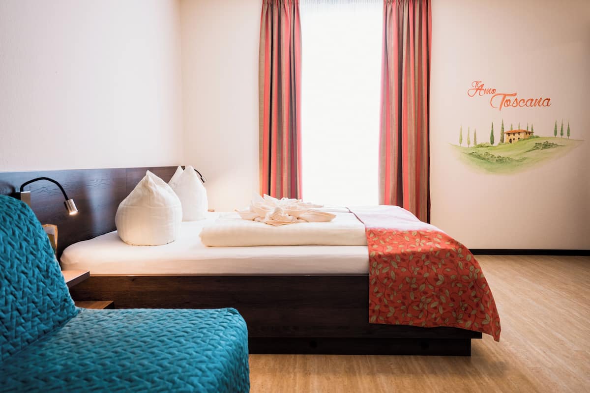 A neatly made bed with a red floral bedspread, a white pillow, and a white throw blanket. There is a blue knitted chair to the left of the bed. The room has a wooden floor and a wall with a mural of a house and trees.
