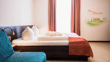 A neatly made bed with a red floral bedspread, a white pillow, and a white throw blanket. There is a blue knitted chair to the left of the bed. The room has a wooden floor and a wall with a mural of a house and trees.
