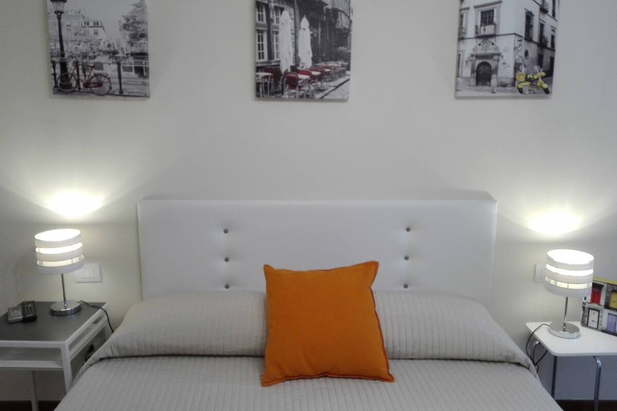 Une chambre d’hôtel bien rangée, avec un grand lit, deux tables de chevet dotées de lampes et trois photographies en noir et blanc accrochées au mur.