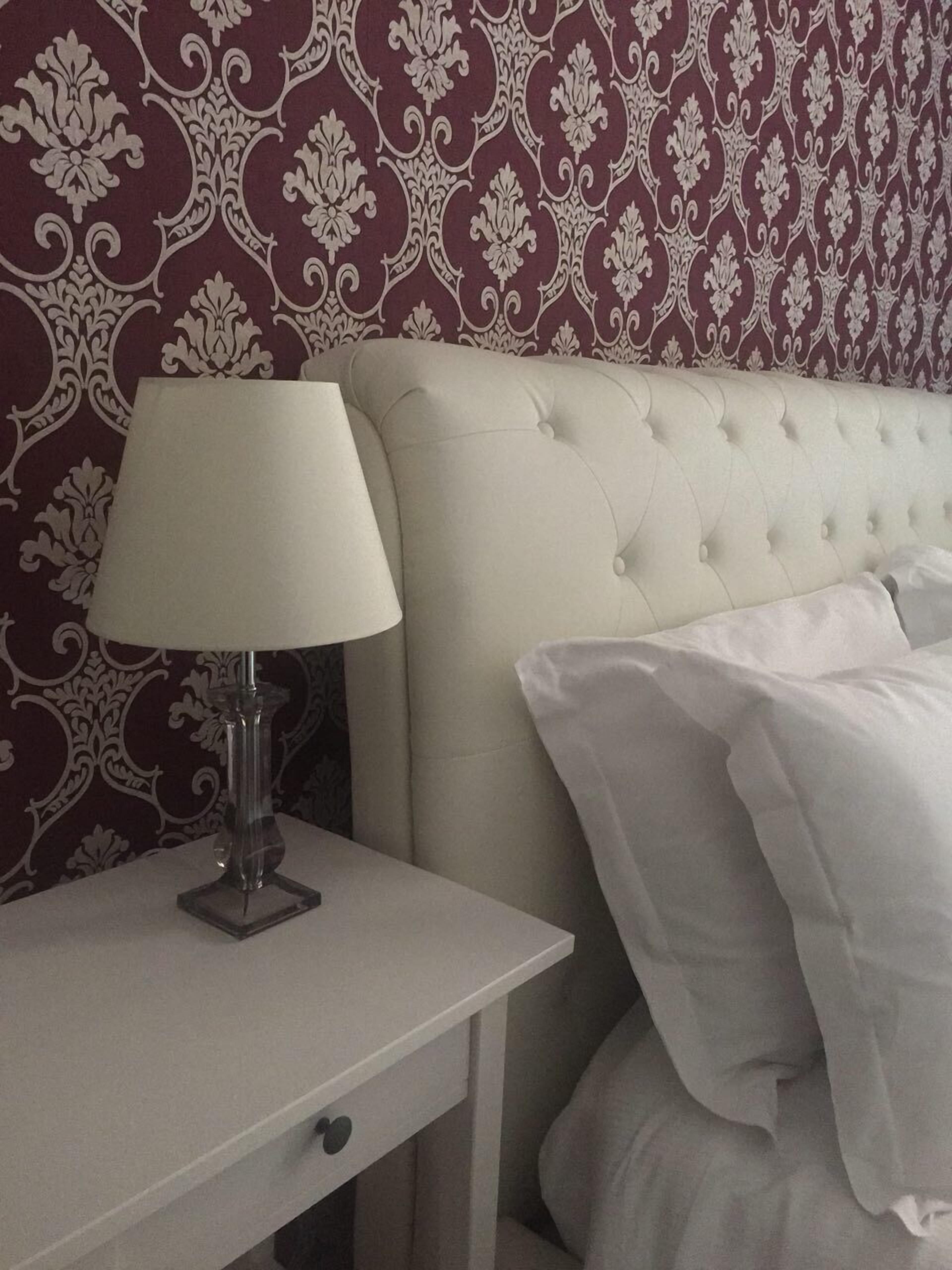 Cama bem arrumada com roupa branca, mesa de cabeceira branca com abajur, e parede com papel de parede estampado.