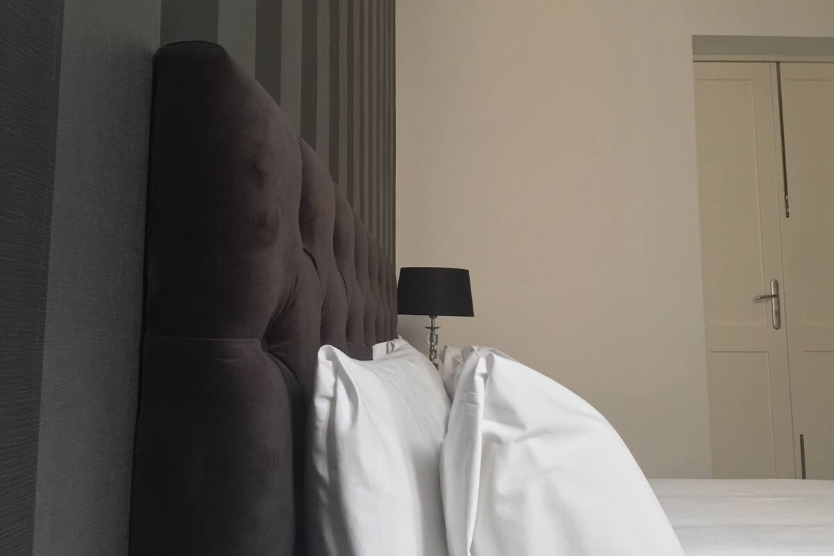 Ein Hotelzimmer mit hohem, gepolstertem Kopfteil, einer Nachttischlampe und einer geschlossenen Tür.