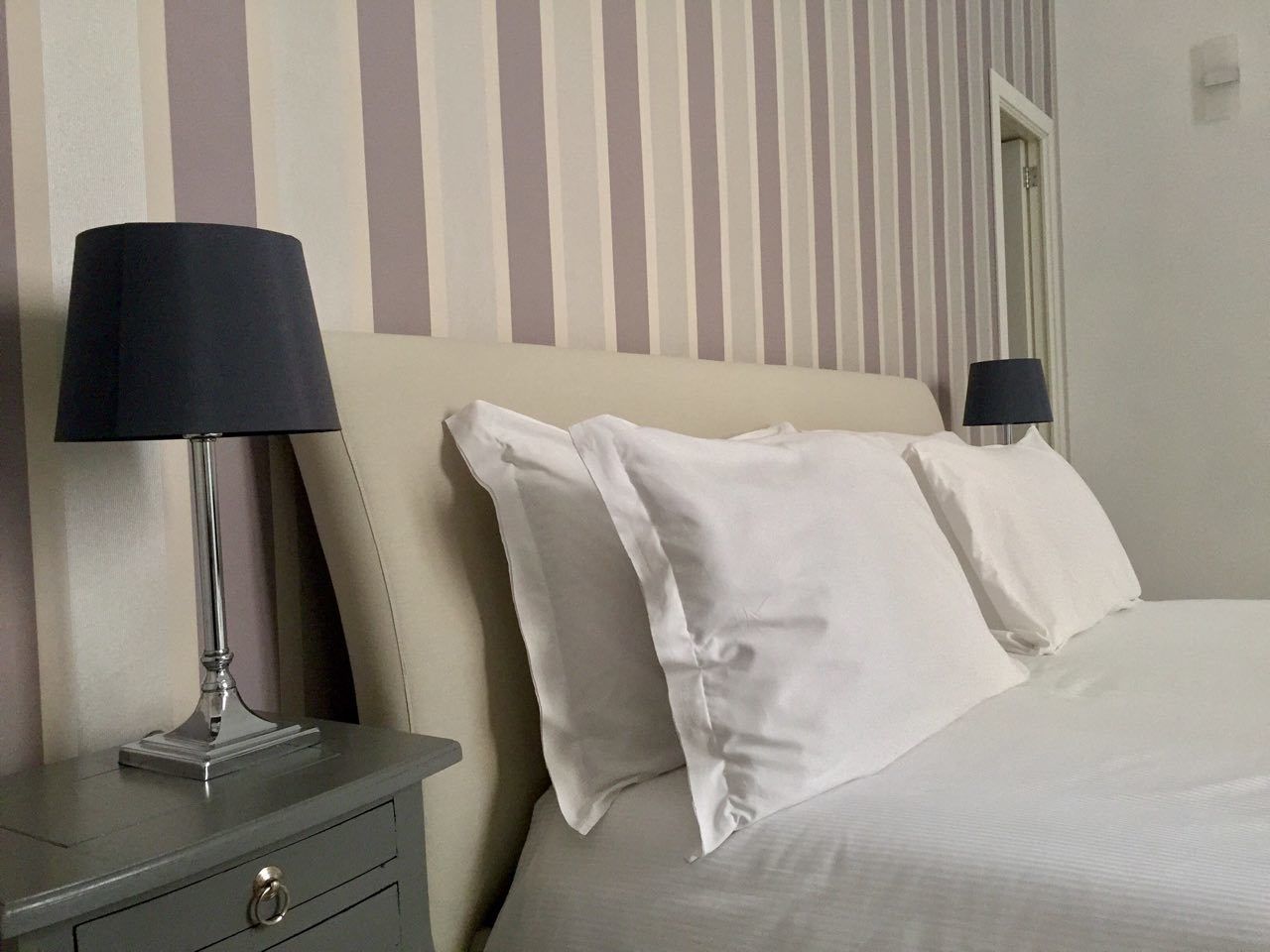 Una cama bien tendida con sábanas y almohadas blancas, una mesita de noche con lámpara, y una pared a rayas al fondo.