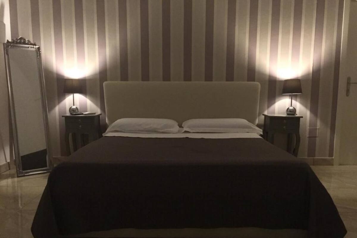 Ein ordentlich bezogenes Bett mit dunkler Tagesdecke und zwei Kissen, daneben zwei Nachttische mit Lampen, in einem Zimmer mit Streifenmuster an der Wand.
