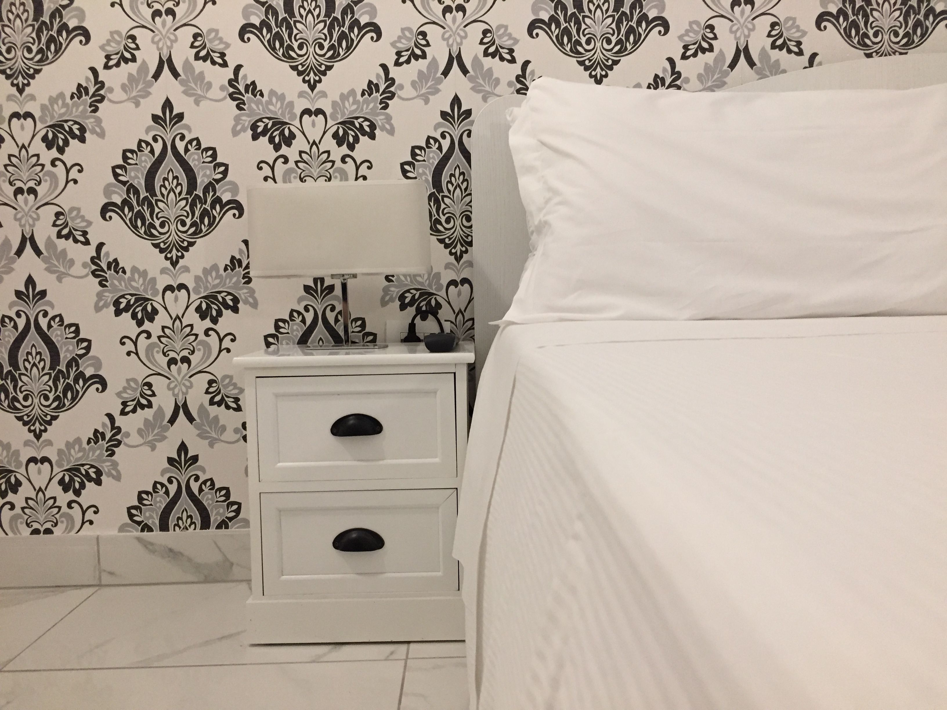 Un dormitorio con una cama blanca, una mesita de noche con dos cajones y un papel tapiz estampado.