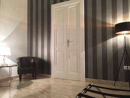 Una habitación con papel tapiz a rayas, una puerta blanca, un sillón de cuero negro, una mesita con una lámpara y una lámpara de pie.