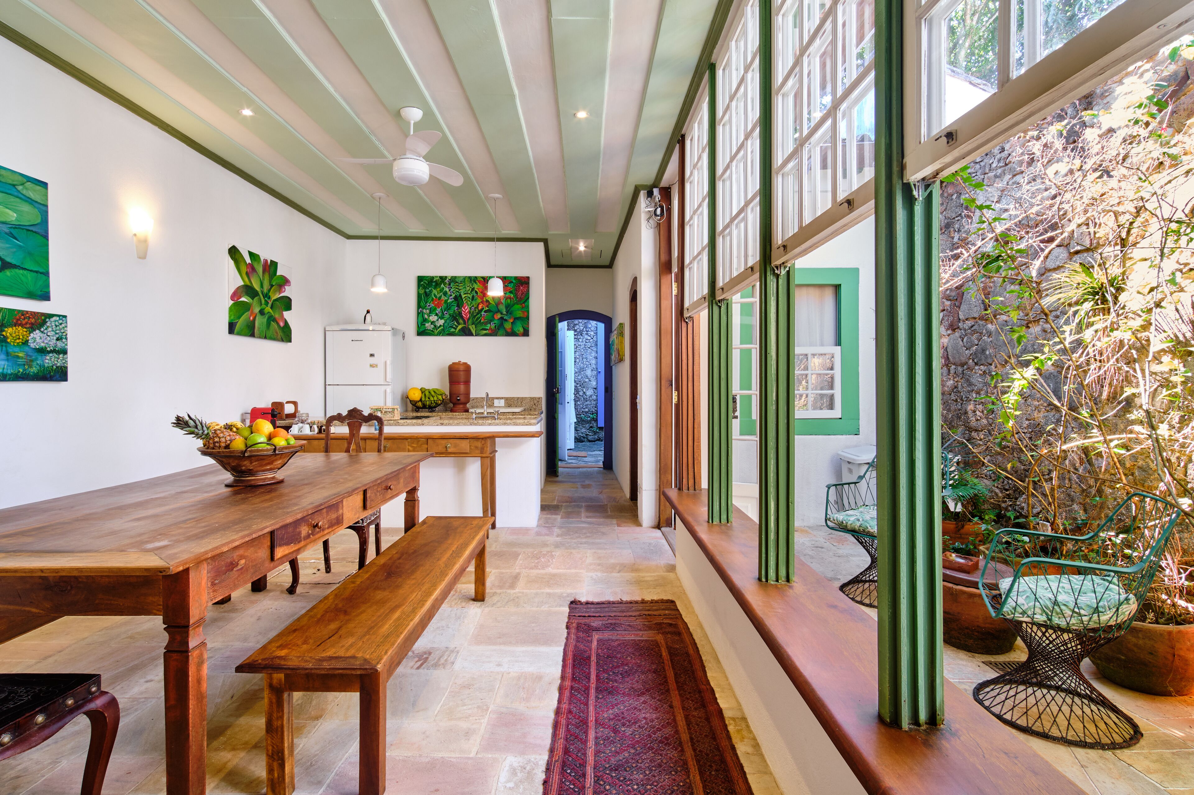 A dining area with a wooden table, benches, and a fruit bowl. There are potted plants and a view of a garden through large windows.