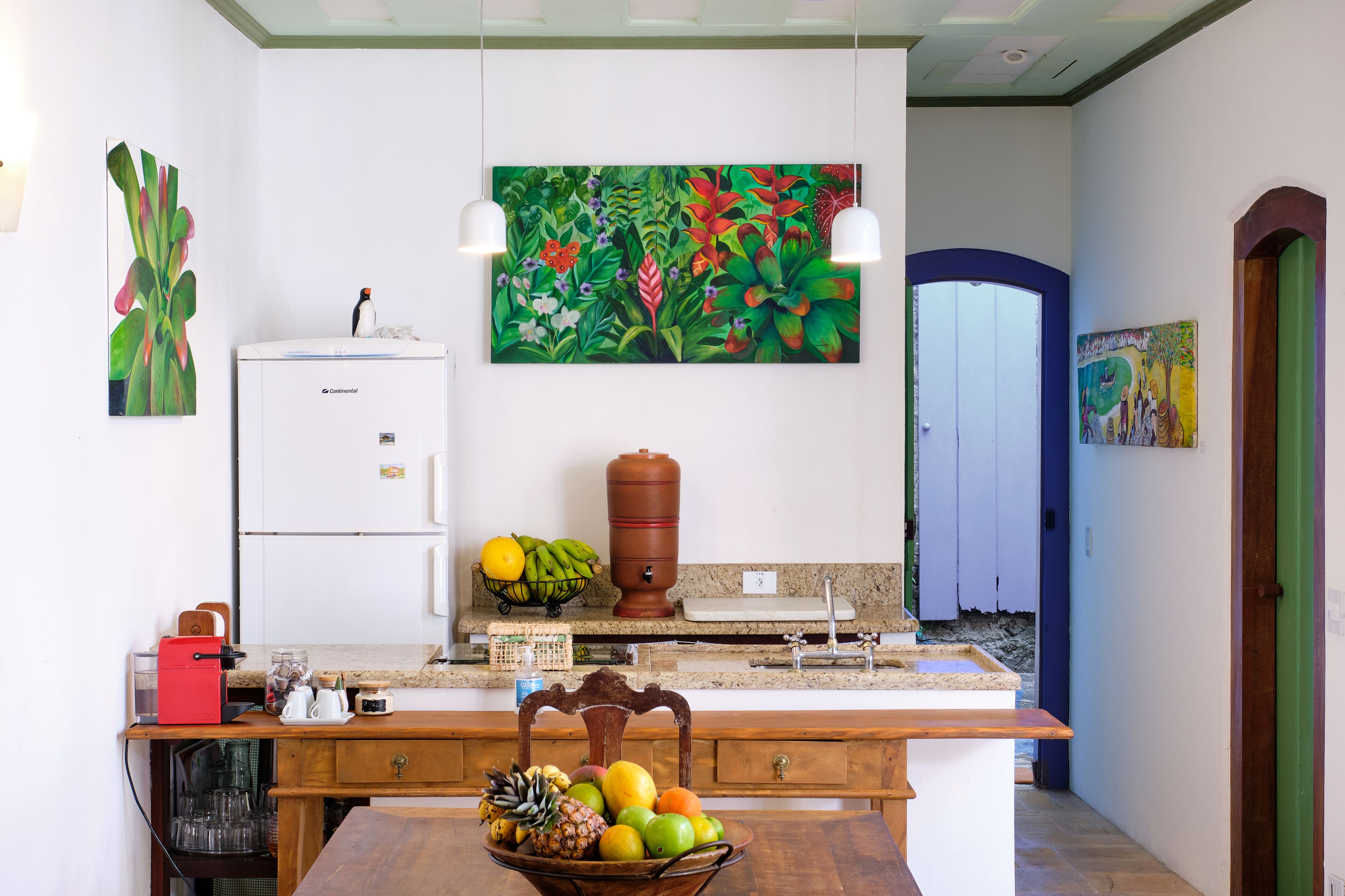 Una cocina con refrigerador, cafetera, frutero y un cuadro en la pared.
