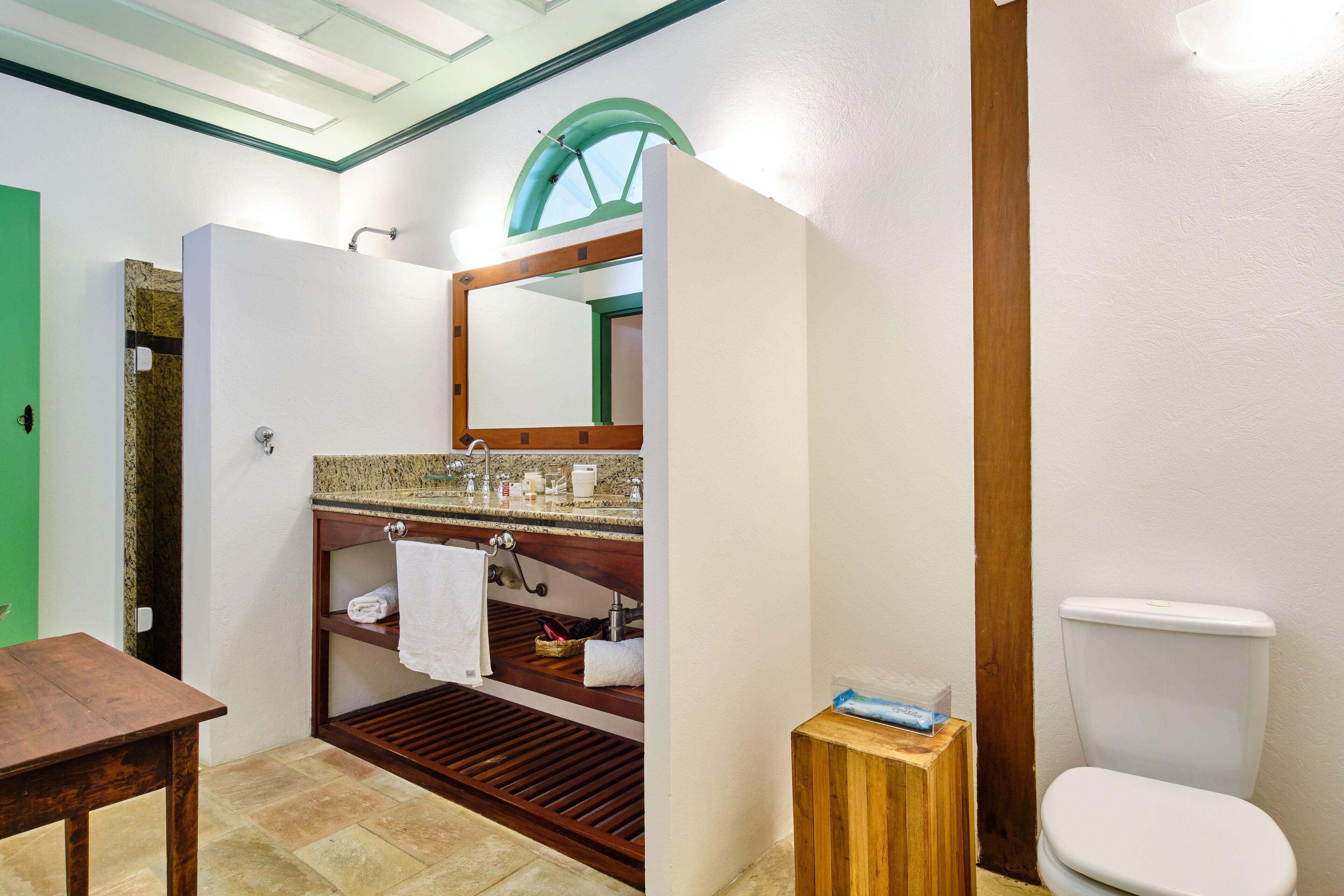 Un baño con tocador de madera, una puerta verde, inodoro blanco y un taburete pequeño de madera.