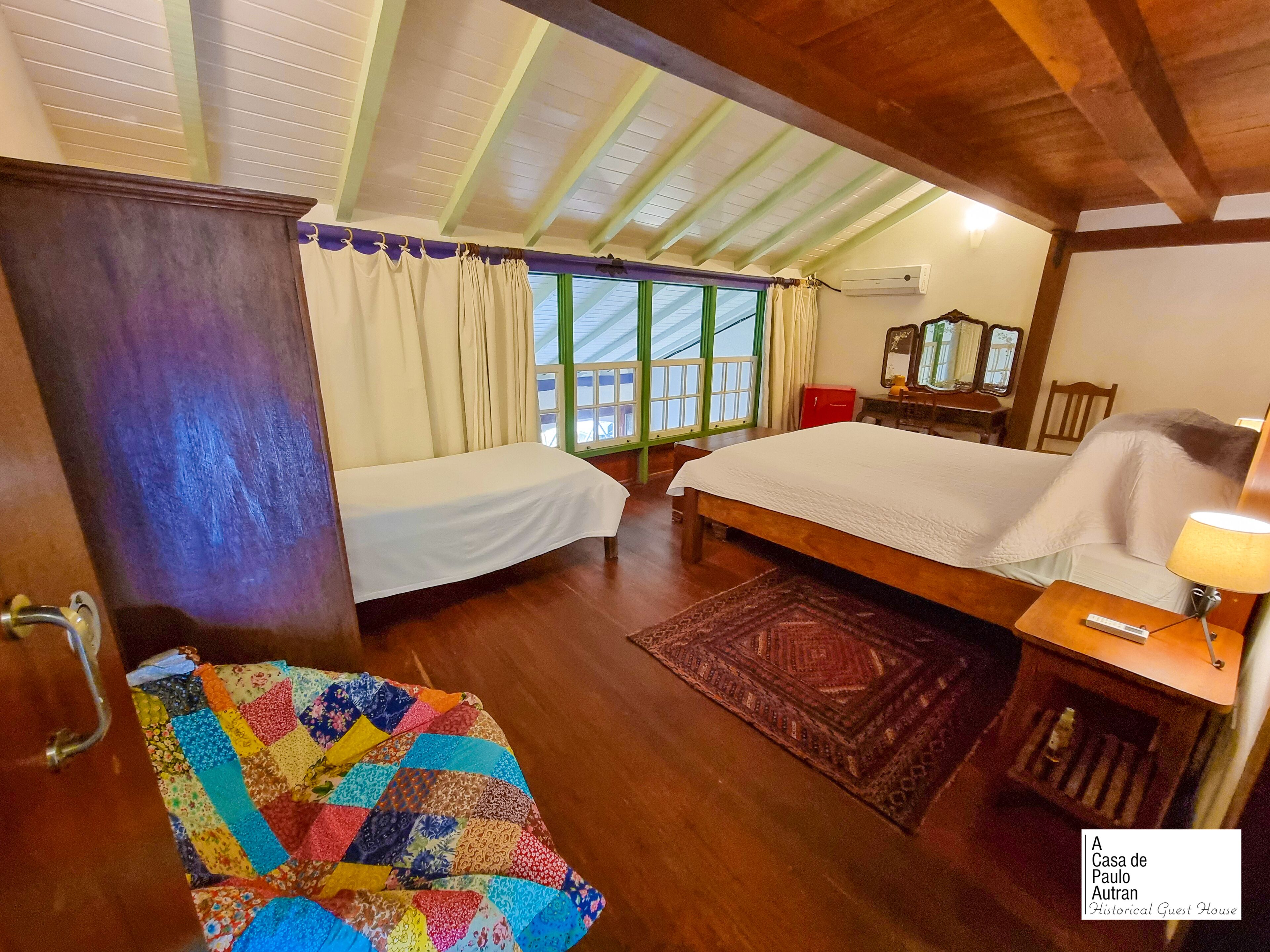 A bedroom with a large bed, a smaller bed, a wooden wardrobe, a mirror, a bedside table with a lamp, and a colorful quilt on the floor.