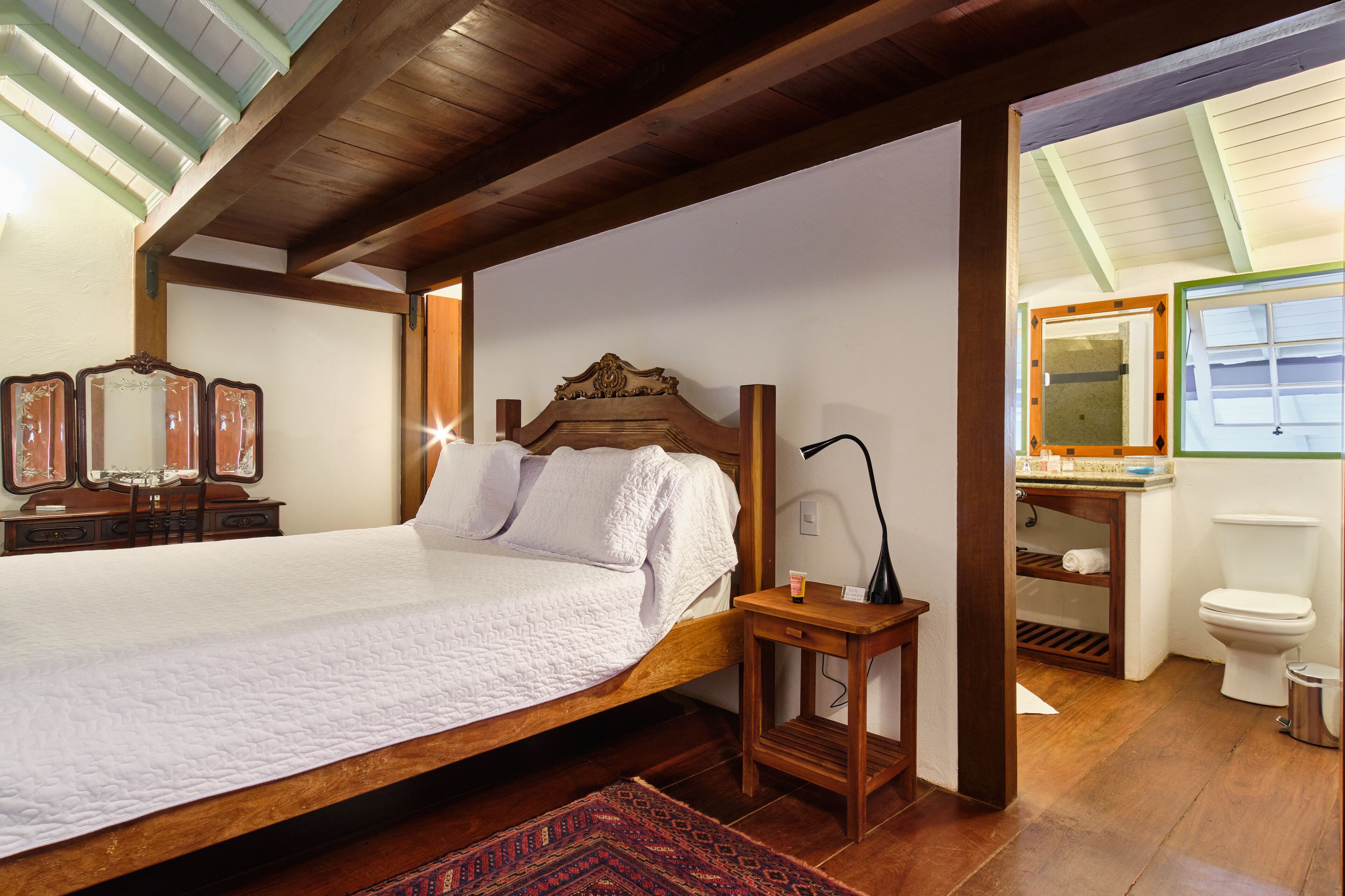 Een slaapkamer met een houten bed, een nachtkastje, een spiegel, en een badkamer met toilet en wastafel.
