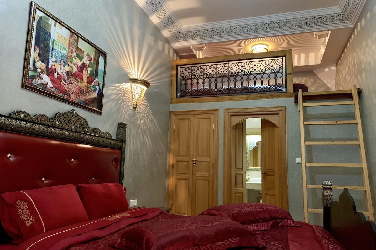 Ein Schlafzimmer mit einem rot gepolsterten Kopfteil, einem Bild an der Wand, einer dekorativen Leuchte und einer Holzleiter.