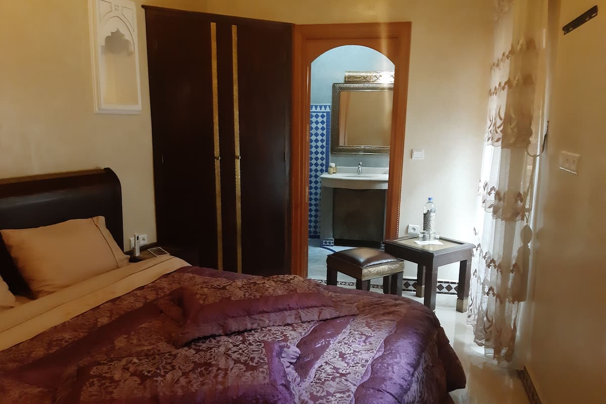 Ein Schlafzimmer mit einem großen Bett, einem Holzschrank, einem Schreibtisch mit Stuhl und einem durchgehenden Türspalt einsehbarer Badezimmer.