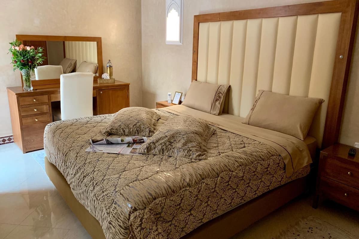 Ein Schlafzimmer mit einem großen Bett, einem Holz-Kopfstück, einem Nachttisch, einer Kommode, einem Spiegel und einer Vase mit Blumen.