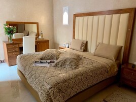 Un dormitorio con una cama grande, cabecera de madera, mesita de noche, cómoda, espejo y un florero con flores.