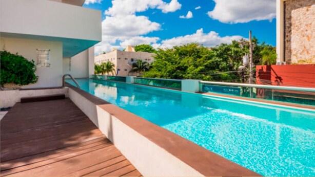 Un hotel moderno con piscina, terraza de madera y vistas a la vegetación y edificios circundantes.