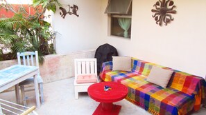Un petit espace de vie extérieur, à l’abri, comprenant un canapé blanc, une table rouge, une chaise blanche et une petite table. On y trouve également des plantes et une fenêtre avec un rideau.