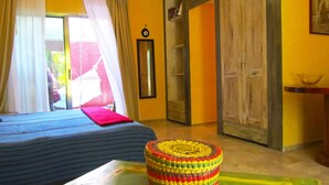 Una habitación con una cama, un taburete tejido de colores, un espejo y un armario.