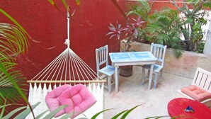 Un pequeño patio con mesa y sillas blancas, un colgador de luces y varias macetas con plantas.