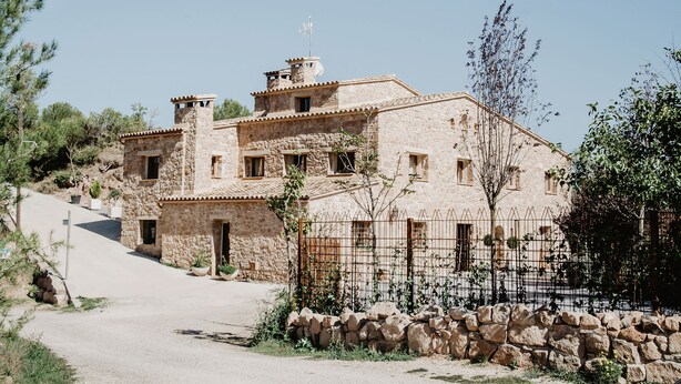 A stone building with multiple windows and a small balcony, surrounded by a stone wall and a metal fence. - Vila Caelus Masia Boutique (Rubió)