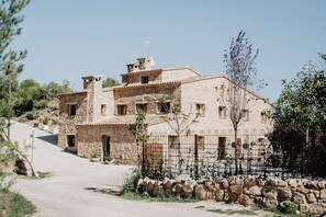 A stone building with multiple windows and a small balcony, surrounded by a stone wall and a metal fence. - Vila Caelus Masia Boutique (Rubió)