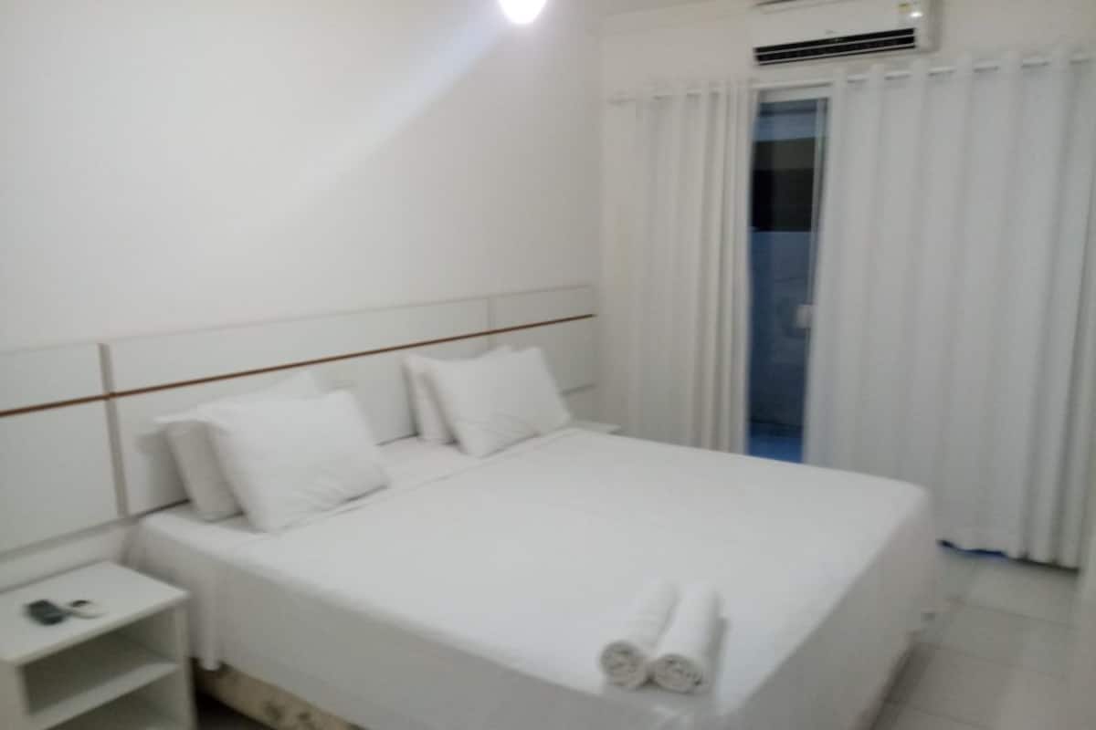 A neatly made bed with white linens and two rolled towels at the foot.