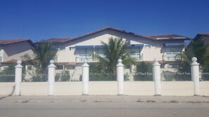 A row of white buildings with balconies, palm trees, and a clear blue sky. - Condomínio Residencial Monte Sião (Porto Seguro)