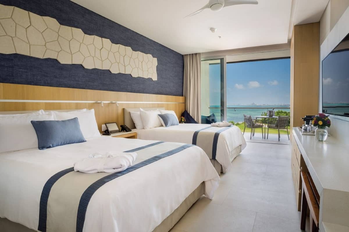 A hotel room with two beds, a balcony with a view of the ocean, a desk with a chair, and a large window.