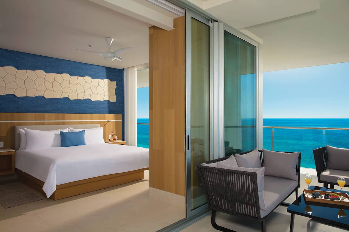 A modern hotel room with a large bed, a balcony with a view of the ocean, and a seating area with a table.