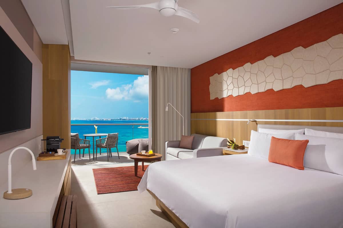 A hotel room with a large bed, a desk, a chair, a small table, a sofa, a TV, and a view of the sea.