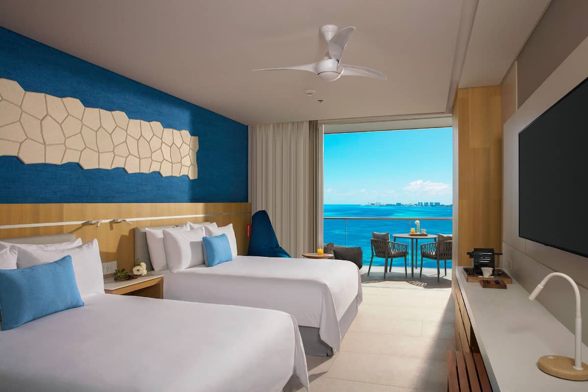 A hotel room with two beds, a ceiling fan, a large window with a view of the sea, a small table with chairs, and a TV on the wall.