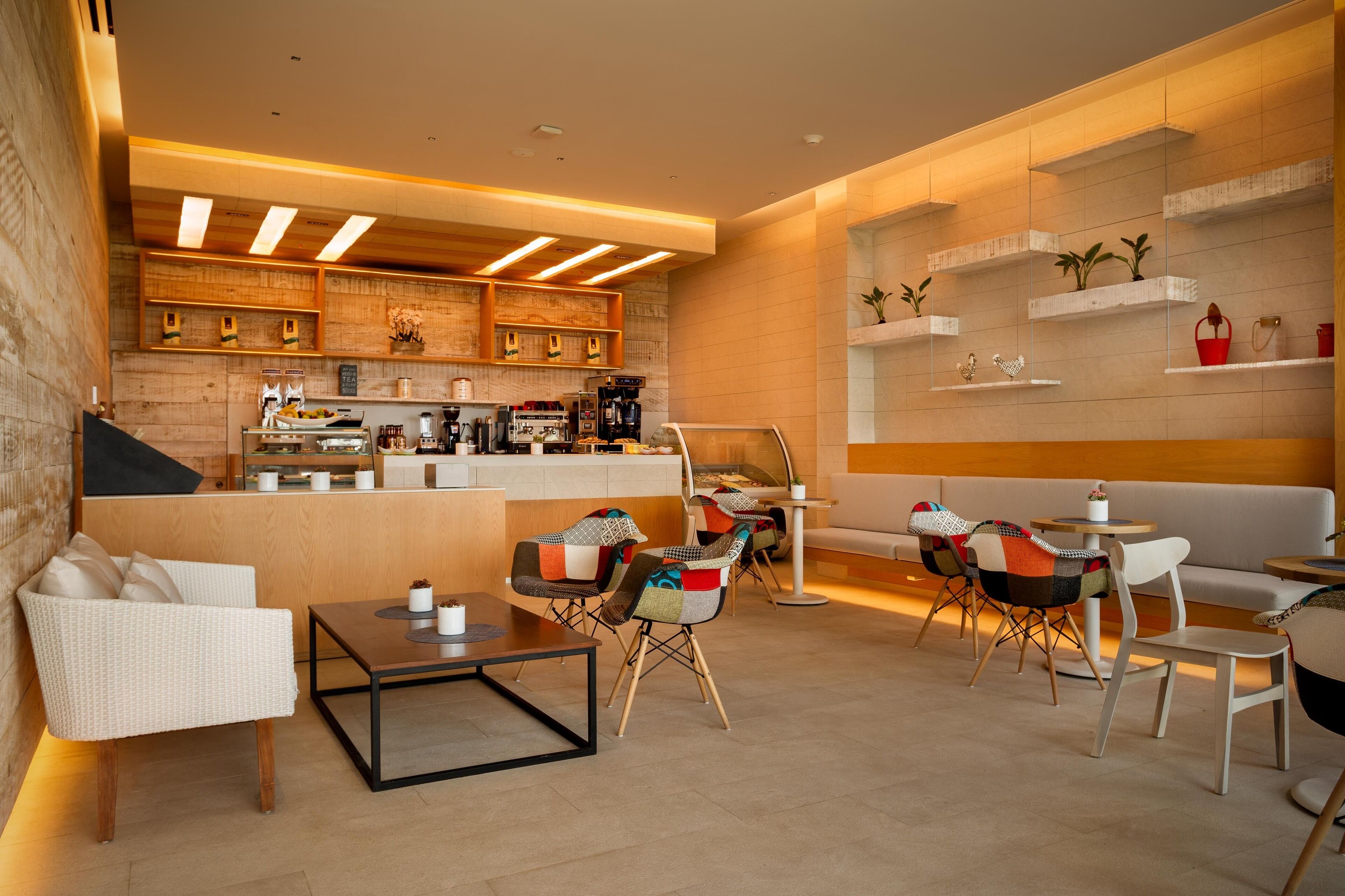 Un café moderno con mostrador de madera, zona de estar con sillas y mesas, y estanterías con plantas y objetos decorativos.