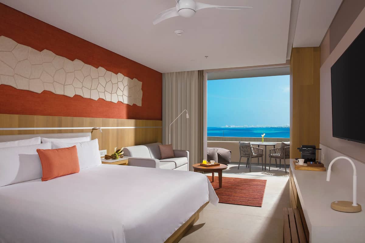 A hotel room with a large bed, a sofa, a small table, and a balcony with a view of the ocean.
