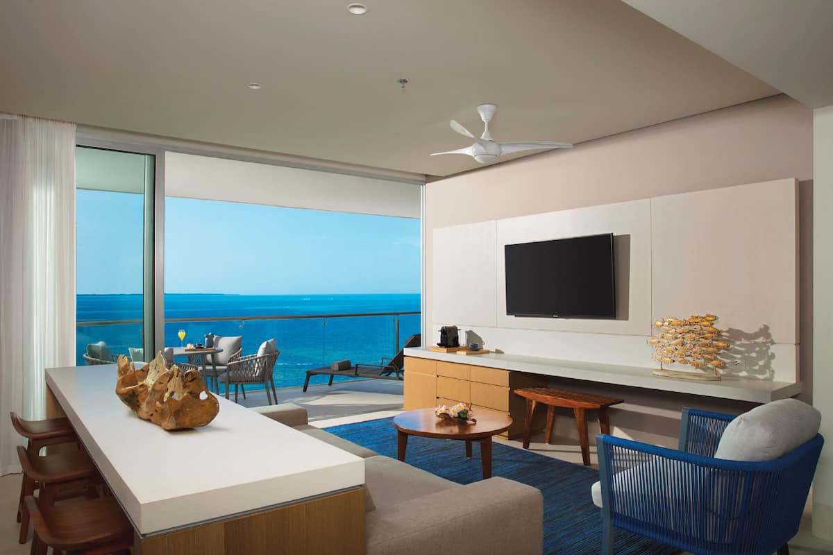 A modern hotel room with a large balcony, a flat-screen TV, a wooden coffee table, and a blue chair.