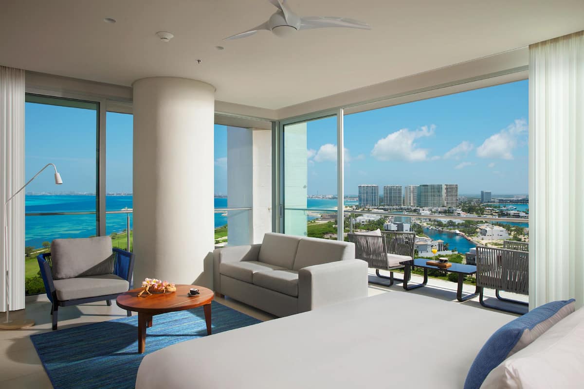 A spacious hotel room with a large bed, a sofa, a chair, and a small table. The room has large windows offering a view of the ocean and cityscape.