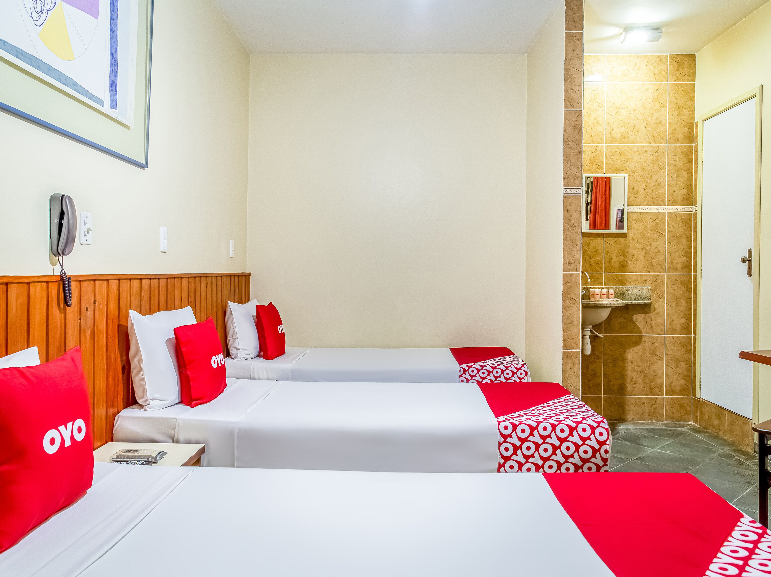 Quarto de hotel com duas camas, cabeceira de madeira, um quadro na parede, um telefone na parede e banheiro com pia e espelho.